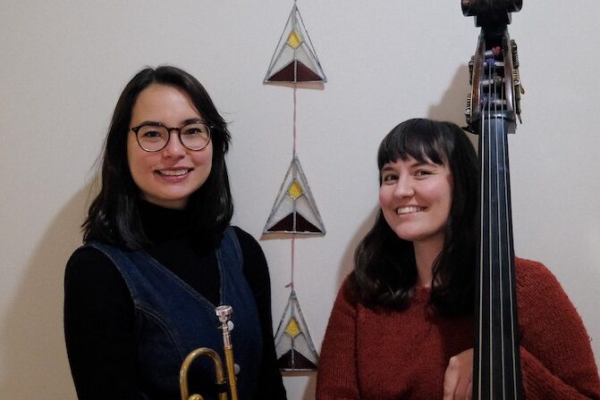 Two dark-haired musicians, one holding a trumpet, the other a bass, smile into the camera. They are Perth jazz artists Jessica Carlton and Kate Pass.