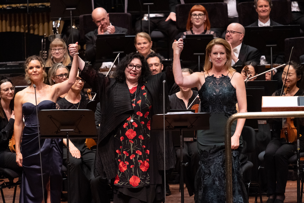 Three elegantly dressed women stand holding hands, arms raised toward an audience, an orchestra smiling behind them. They are singer Sara MacIver, composer Elena Kats-Chernin and singer Emma Campbell.