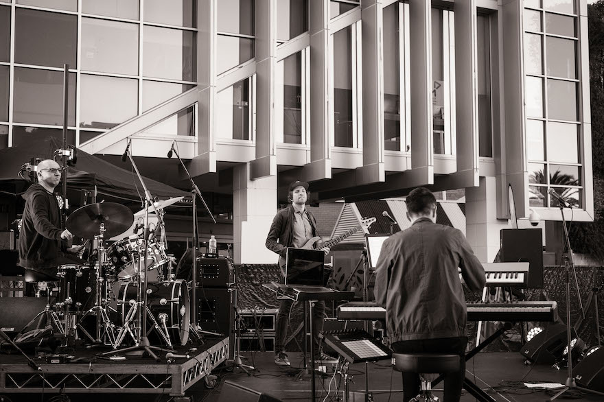 Three musicians are spread across a stage in front of tall windows covered in column. One is to the left, sitting behind his drum kit, another has his back to the camera facing his keyboard, with the guitarist standing in the middle to the rear. They are Perth trio Trisk.