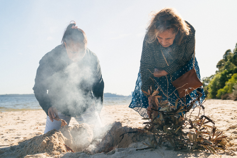 Melville Storylines promotional image: Two women peer down into a hole dug on a sandy beach emanating smoke. The woman on the left is partially obscured by the smoke. The woman on the right is reaching down into a pile of drying leaves.
