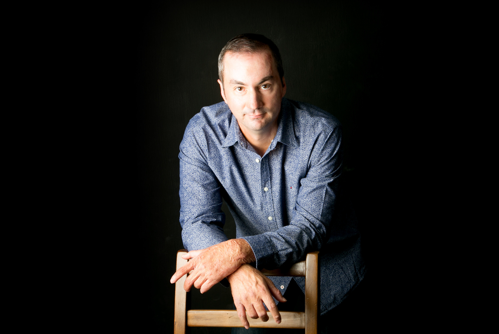 Melville Storylines promotional image: A middle-aged man with close-cropped hair poses against a black studio background, gazing into the camera as he rests his crossed arms against the back of a wooden chair. 