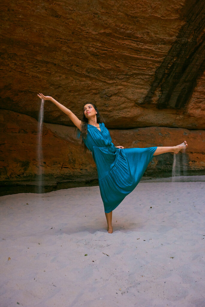 A dancer poses on one leg on the sand in front of red ochre cliffs. The other leg is outstretched, her arm raised with sand falling through her fingers. This is Rika Hamaguchi in Tourism Western Australia's Walking on a Dream campaign. 