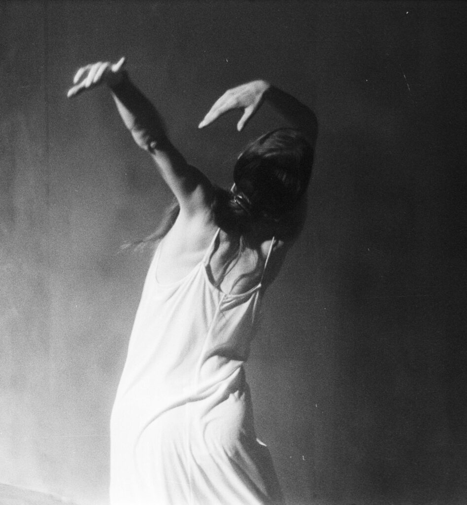 A black and white image of a woman in a slip dress with her back to the camera, her arms tilting back above her head