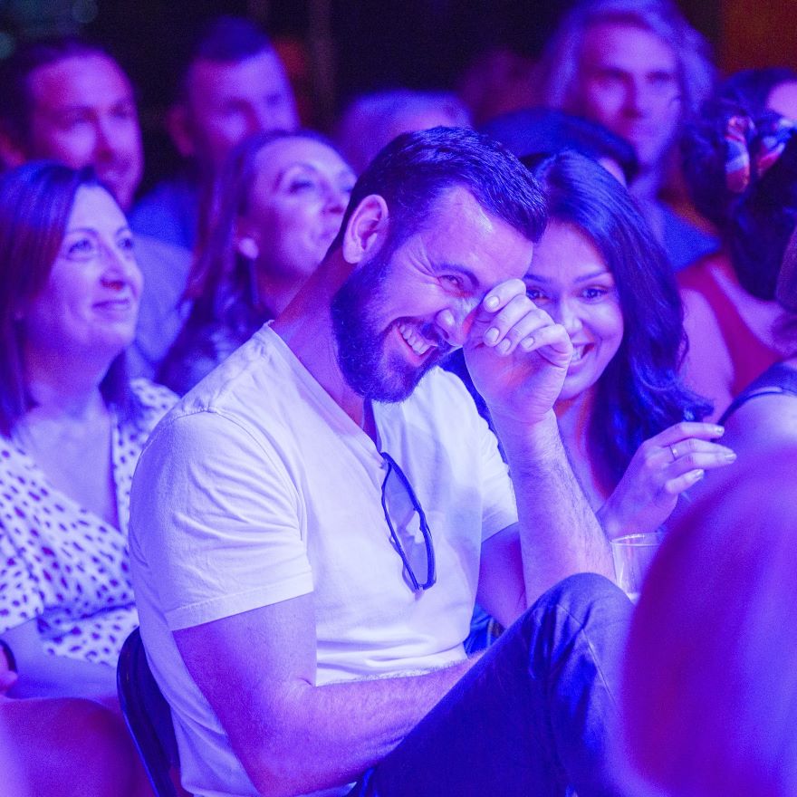 An audience of people smiling, in the foreground a man in a white shirt wipes his eyes as he laughs