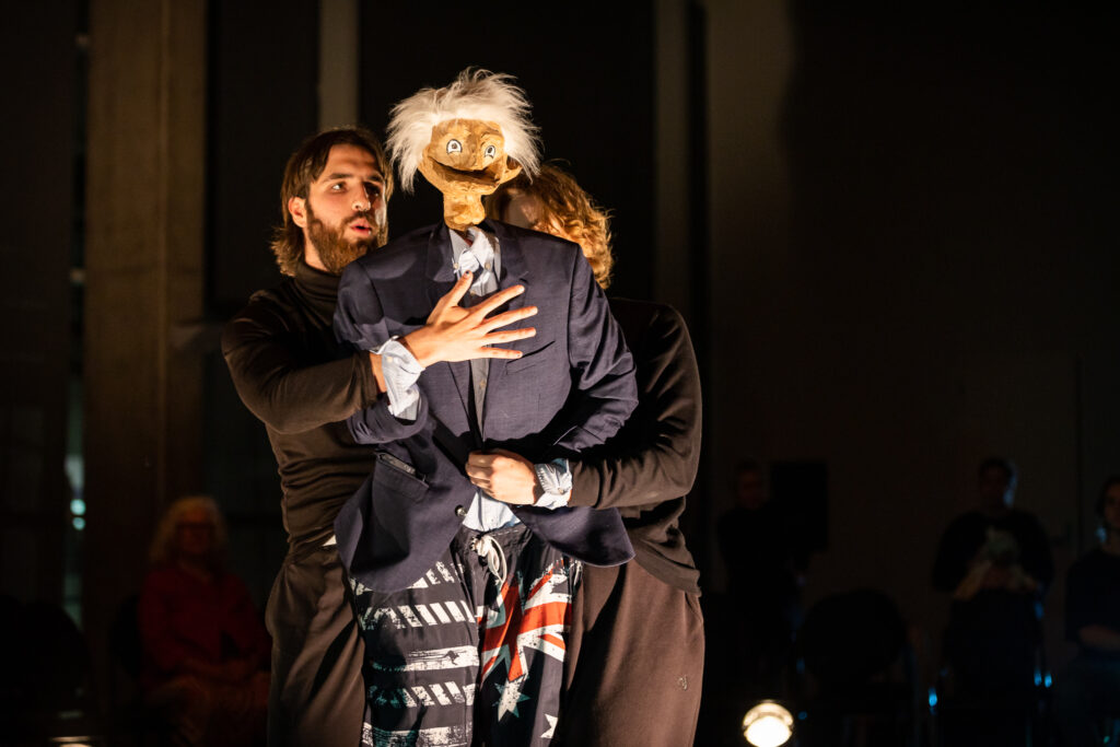 A silver-haired puppet wearing a suit is manipulated by two performers. It is the Premier in WAAPA and Spare Parts Puppet Theatre's The Last Ship Left