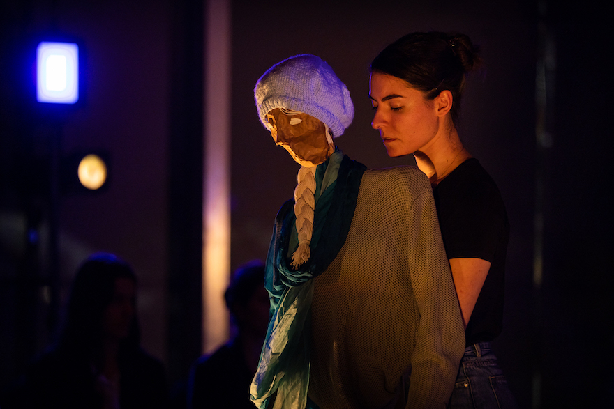 A performer holds a puppet dressed as an older woman, hair in a braid, head cast down. They both look sad. This is a scene from WAAPA and Spare Parts Puppet Theatre's The Last Ship Left.