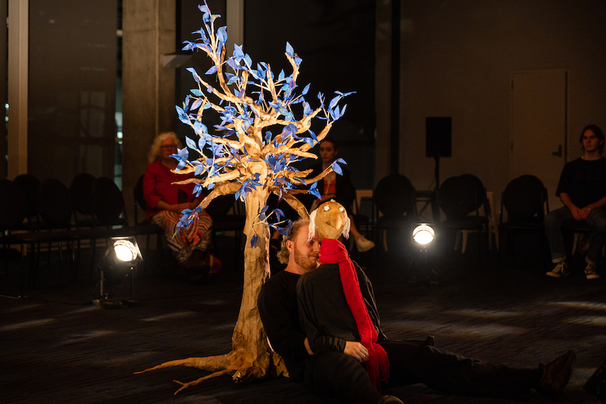 A man holding a puppet sits, leaning on a tree bright with blue lights. An audience sits around the edges of the big room. It is a scene from WAAPA and Spare Parts Puppet Theatre's The Last Ship Left.