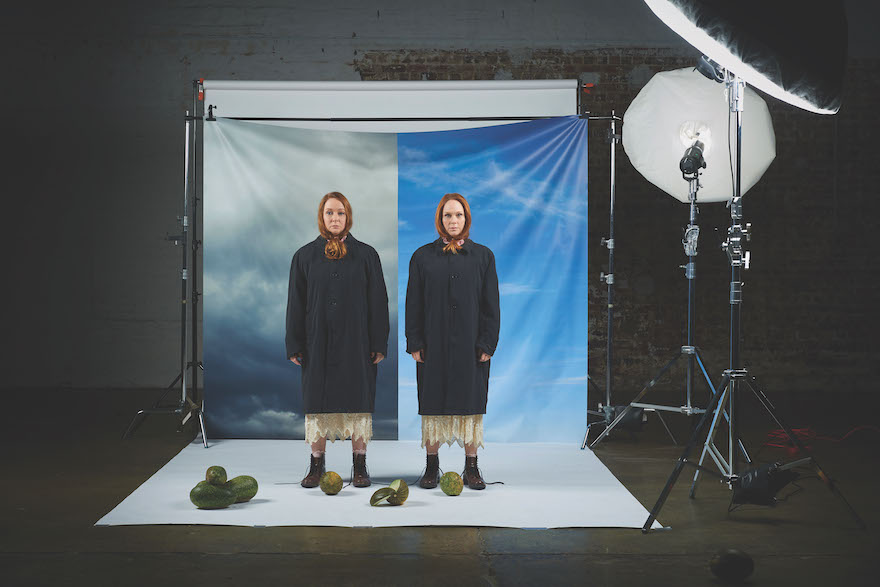 Two red-haired sisters dressed in exactly the same clothes - dark coats, cream skirts and boots - stand in a studio, lighting off to the side. A sheet hangs behind them, one half framing the first woman in a gloomy sky, the other a blue sky. It is Mandy and Hayley McElhinney for 'Dirty Birds'.