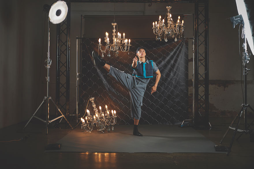 A dancer clad in grey with a blue-edged crop top kicks his right leg high. He is framed by three chandeliers, two above him and one upside down on the ground. There is studio lighting either side of him. This is a photo shoot for Marrugeku's 'Jurrunga Ngan-ga'