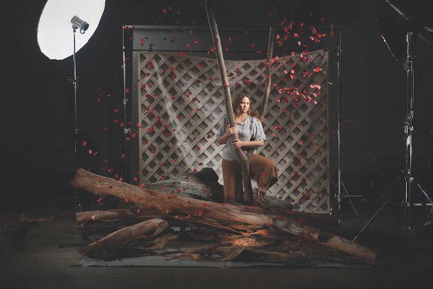 A woman in a grey top and brown pants stands, one leg perched onto a pile of logs, holding onto a v-shaped branch. There are reddish petals all around her. This is a studio shoot for The Blue Room's The Bleeding Tree.