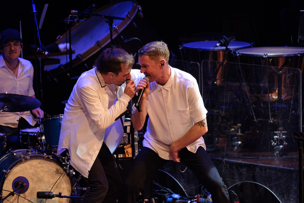 Two singers dressed in white tops and dark pants, lean into each other, smiling as they sing into microphones.  They are surrounded by drums and other percussion instruments, a drummer can be seen behind them. This is Morgan Bain and Drapht performing with WASO.