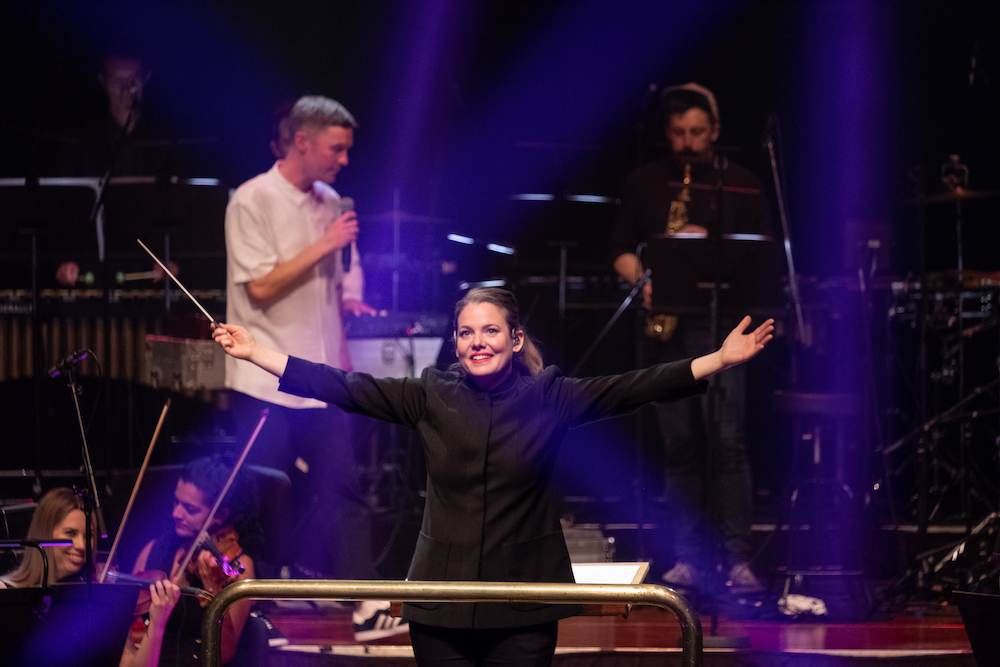 A woman in a long-sleeved black top stands arms outstretched, her right hand holding a baton. Behind her a singer in white top holds a microphone, and musicians perform on strings and brass. This is WASO assistant conductor Jen Winley with hip-hop artist Drapht.