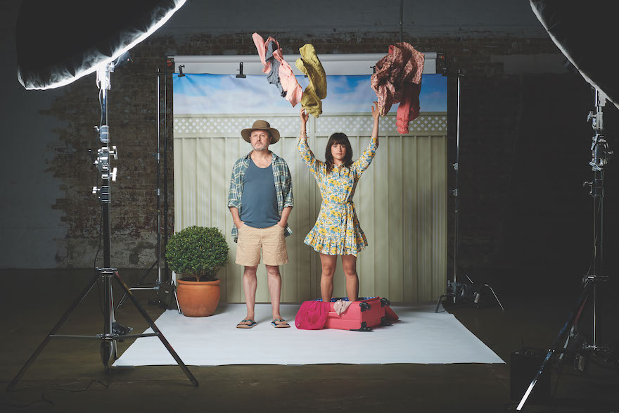 A couple stand under studio lights, against the backdrop of a suburban fence, a potted plant off to one side. The man is wearing thongs, shorts, and a checked shirt over a singlet, a hat on his head. The woman, wearing a short floral dress, is standing in an open pink suitcase, clothes spilling over the edge. She is throwing a bunch of others in the air above her. This is a studio shoot for 'Things I Know to Be True'.