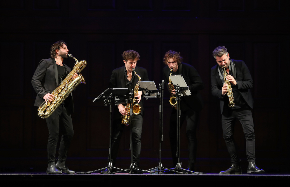 Four musicians stand on stage behind lecterns. They are dressed in black and playing saxophones in an ensemble formation. This is Signum Saxophone Quartet.