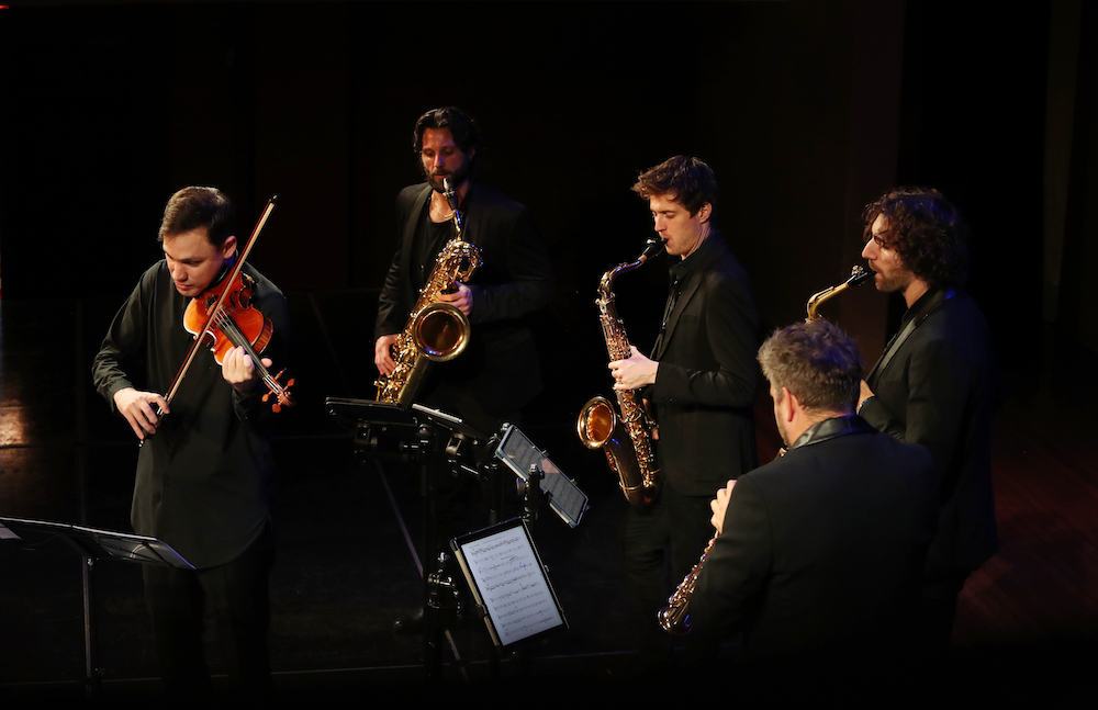 Five musicians clad in black form a semi-circle. To the left, a violinist leans into his instrument, four men on sax around him. These are violinist Kristian Winther and the Signum Saxophone Quartet.