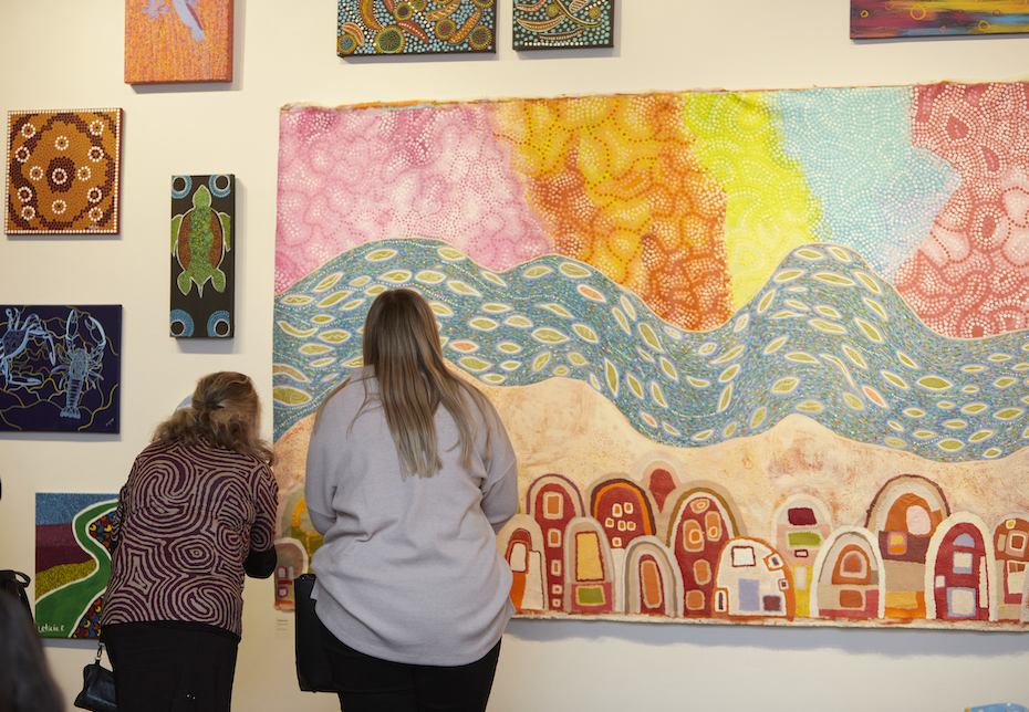 Two people stand in front of a large vivid canvas, their backs to the camera. Smaller artworks by Noongar artists are dotted around the main piece. This is the Noongar Country exhibition that was held at BRAG.