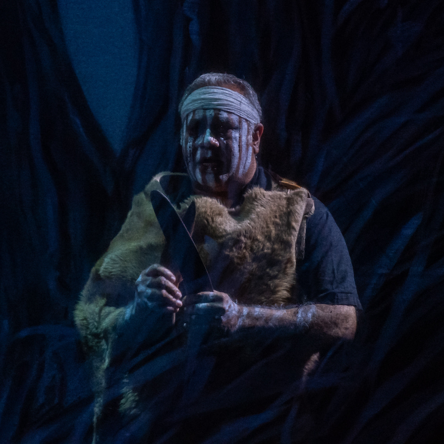An Indigenous man wearing face paint and a traditional cloak looks plaintive, his hands out in front of him as if trying to say something.  This is Barry McGuire in Swan Lake.