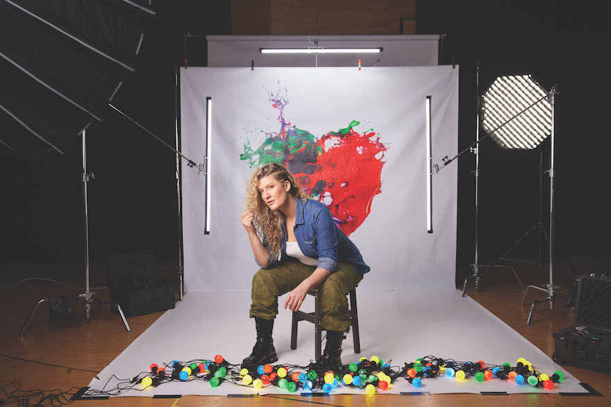 A woman with long blonde hair sits in front of a long white canvas with a red and green splattered heart on it, a string of coloured lights at her feet. There is a stage light to her left and other camera gear off to each side. This is Virginia Gay in Cyrano.