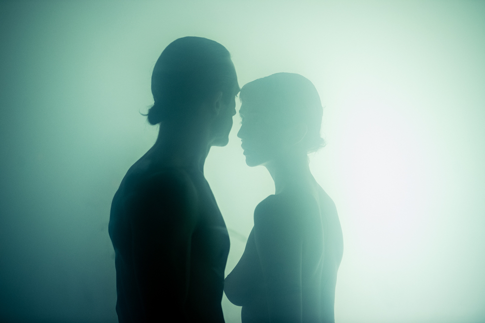 A scene from IN.HOUSE Studio Season in which two people stand opposite each other. We can only see them from the chest up, they are unclothed and swathed in haze.