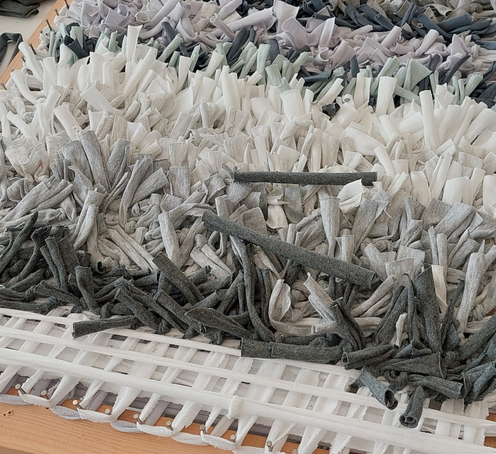 A work from Foraging in the Wastestream - pieces of grey and white scrap fabric are woven through a plastic mesh to form a carpet-like surface,