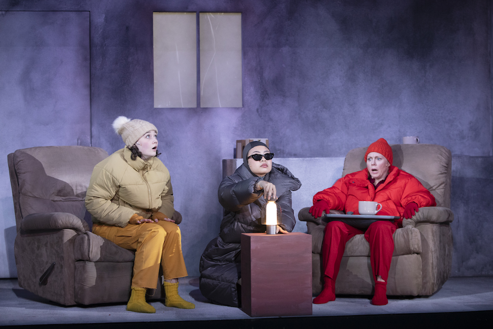 Three people in a grey room sit, dressed in snow gear as if they are freezing. The one in the centre has her  hand above a single light bulb, looking up. The yellow-clad women to her right is also looking up, while the third woman, dressed in red from head to toe, is nursing a tray with a large soup mug on it, has a horrified expression on her face. This is a scene from the Ella Hickson play 'Oil'.