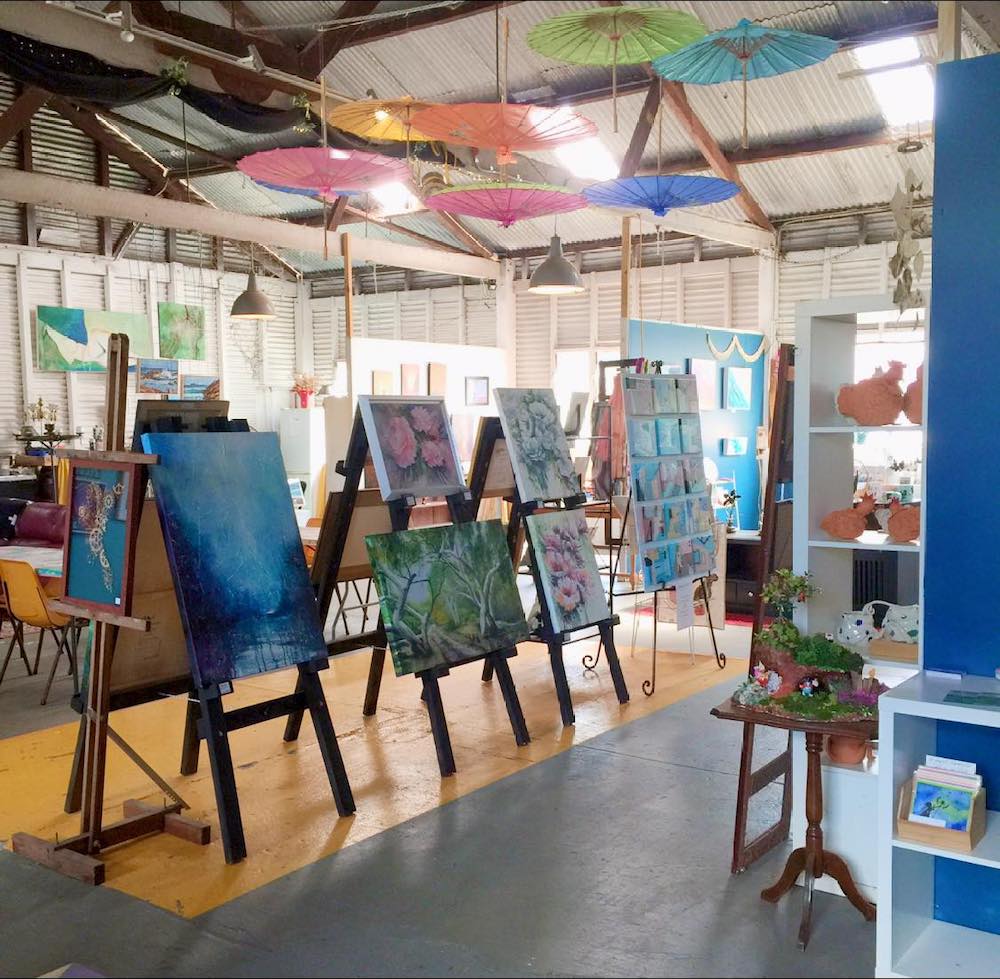 A large light-filled room with high ceilings is filled with colourful paintings arranged on easels. Brightly coloured Asian-style umbrellas hang from the roof, ceramic pieces on the shelves. This is the art hub The Rabbit Hole.