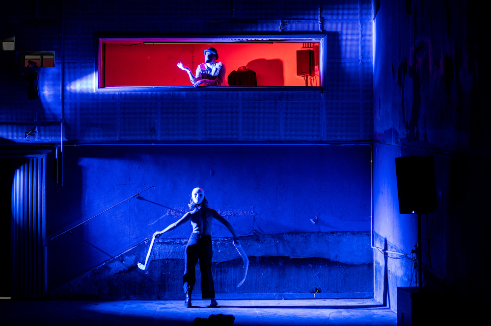 A woman stands with her back to the camera holding what appear to be prosthetic limbs. She is looking up at a man in a long, narrow cinema booth. The walls are cast in blue light, the booth in red. They are Antonio Rinaldi and Celina Hage from STRUT Dance.