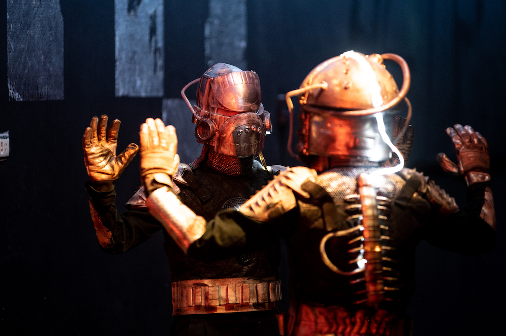 Aliens clad in metallic gold and silver space suits face each other, arms raised, hands up, mirroring each other's movements. They are Georgia Van Gils and Zendra Giraud from STRUT Dance.