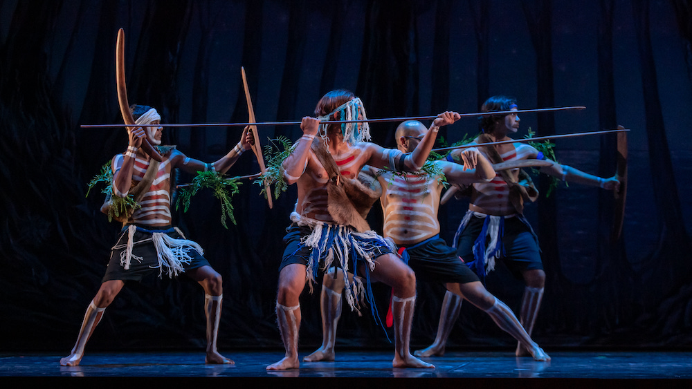 Four Indigenous men, their bodies and faces painted, stand as one, holding what look like spears and boomerangs. They are looking in the same direction, as if facing down an enemy. This is dance group Gya Goop Keeninyarra in Swan Lake.