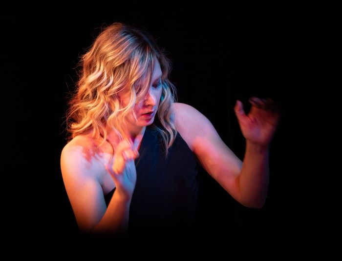 An ink black background from which emerges the head and shoulders of woman with blonde hair, her arms raised as though she is conducting a band.