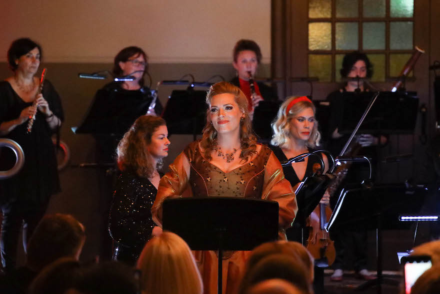 A woman clad in an orange gown, hair elegantly arranged, stands at a lectern, smiling. Behind her a group of musicians holding various instruments concentrate. This is soprano Rachelle Durkin with members of Perth Symphony Orchestra.
