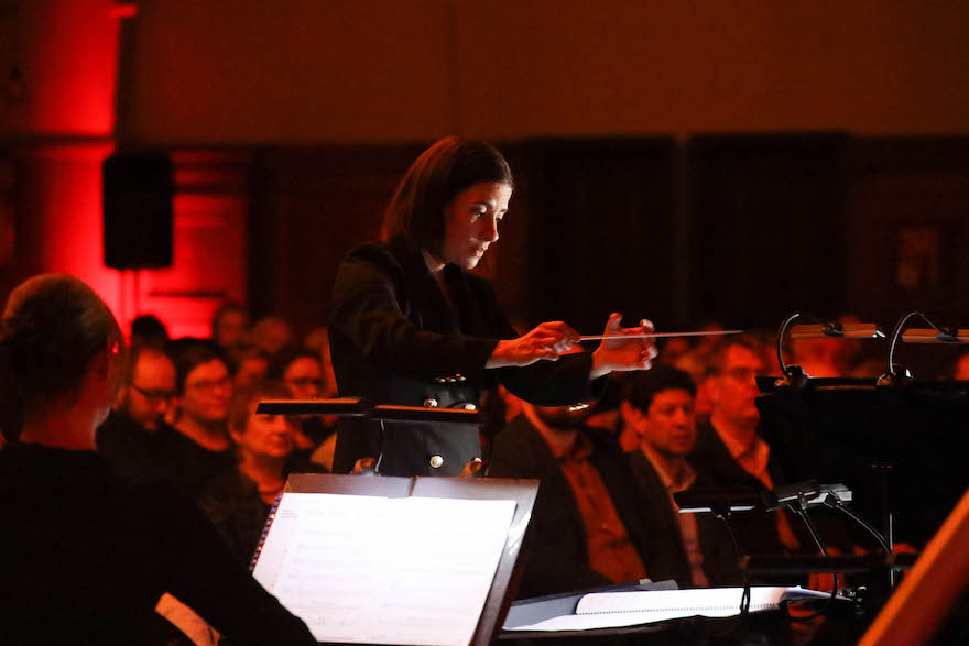 A woman dressed in black has her arms stretched out, one hand holding a baton, facing musicians. An audience cast in red light can be seen in the background. It is Sara Duhig, conducting Perth Symphony Orchestra