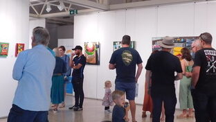 A group of people stand in a light-filled gallery taking in the artwork lining the white walls. This is Collie Art Gallery.