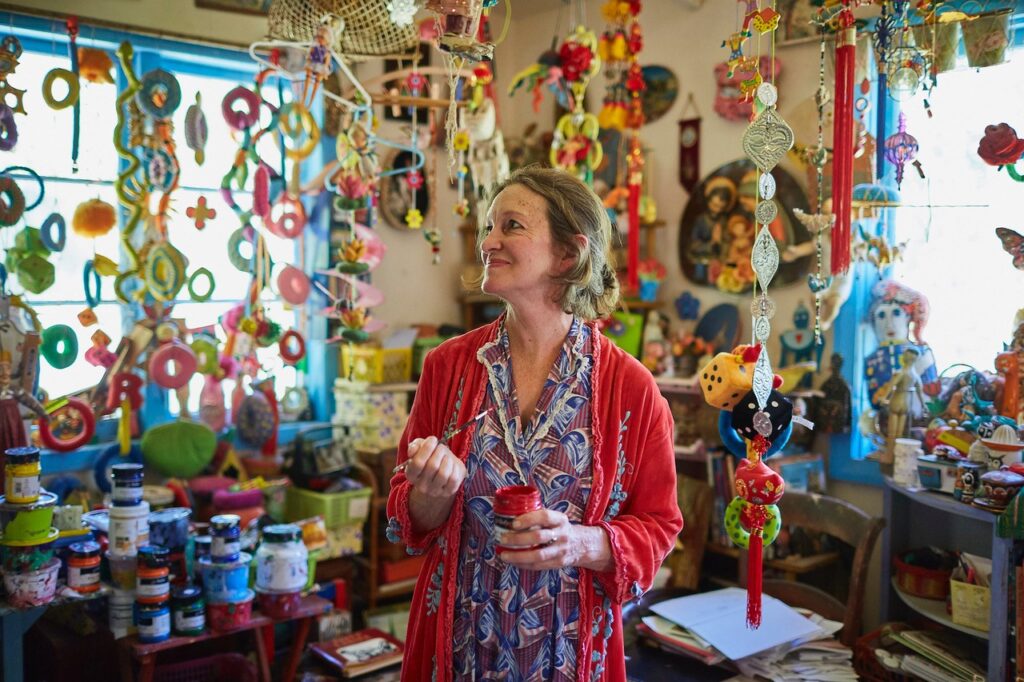 A woman in a patterned dress and red cardigan stands in a studio, paint brush and pot in hand. She is surrounded by colourful creations hanging from the roof, paint pots and other art materials scattered over every surface around her. This is artist Rebecca Cool in her studio for Margaret River Region Open Studios.