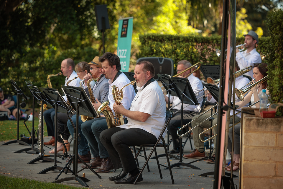 A group of musicians, mostly on trombones and trumpets sit in front of lecterns, playing music. They are in a pretty outdoor setting, with light shining through lush trees. They are part of the Western Australian Jazz Project.