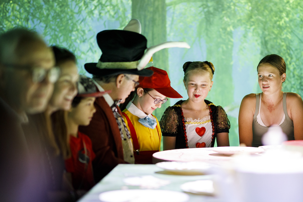 A group of people young and old, some in colourful fancy dress, sit around a table with large plates and cups. Behind them is a magical looking forest scene. This is the tea party room at Wonderland.