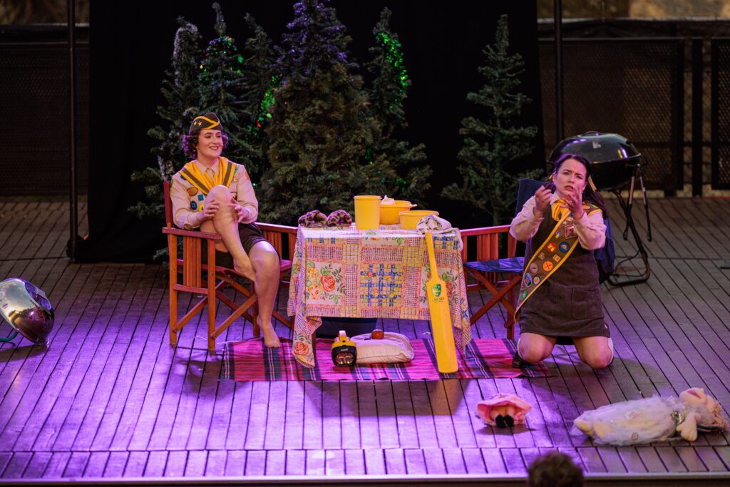 Two young people in Scout-type outfits are outdoors around a table with a cricket bat, flashlight and other camping paraphernalia around them. One is sitting on a chair smiling, a leg tucked up; while the other is kneeling on the wooden deck, arms outstretched. This is Freeze Frame Opera's staging of Hansel and Gretel.