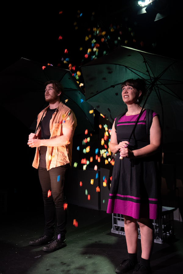 Two people stand under large black umbrellas. Pieces of coloured light or confetti fall from above.