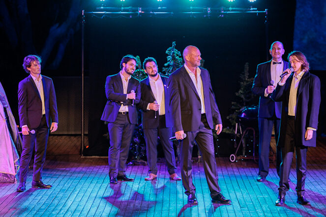 A group of men in dark suits and white shirts stand on a wooden stage, microphones in hand. One is singing into the microphone while the others look on. This is Freeze Frame Opera's Christmas at the Quarry.