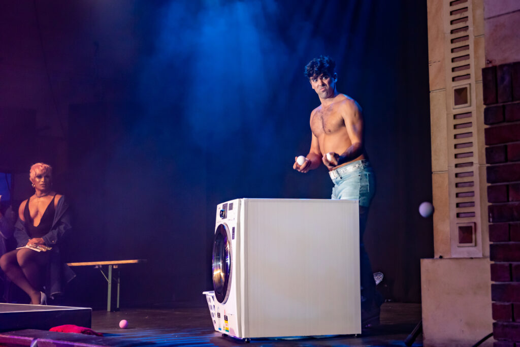 A shirtless person stands behind a washing machine, holding what look like eggs. A drag queen clad in a sexy dress sits on a bench off to the side. 
They are part of cabaret act Breifs appearing at Fringe World.