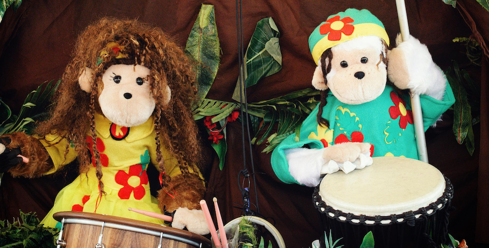 Two soft toy monkeys clad in flowerly outfits, one yellow, the other green, sit in front of drums, a jungle scene behind them.
They are the Amazing Drumming Monkeys, which will be performing at Fringe World.