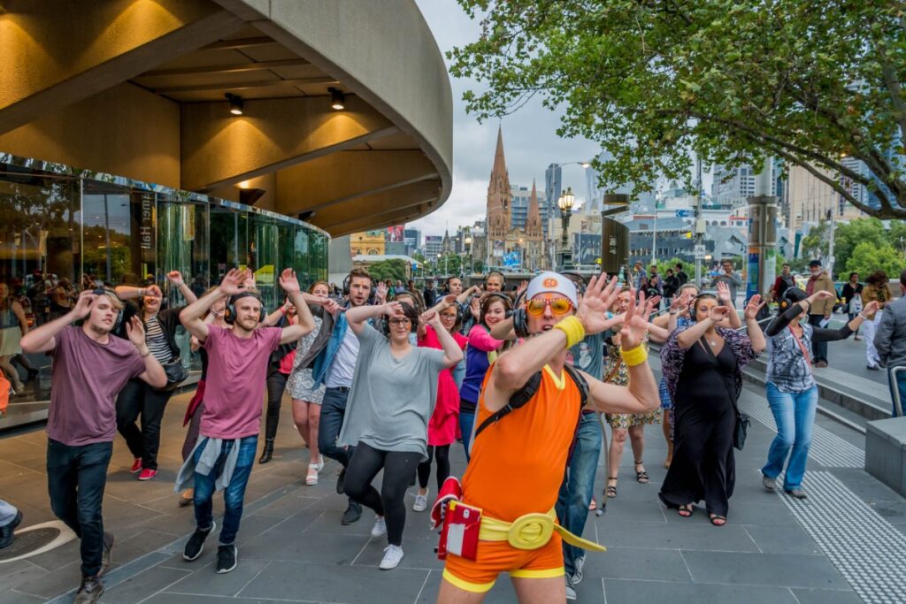 A man in a short orange and yellow jumpsuit and headphones is leading a group of people through a city boardwalk. They are also all wearing headphones and dancing. This one of Guru Dudu's walking tours, which will be part of Fringe World.