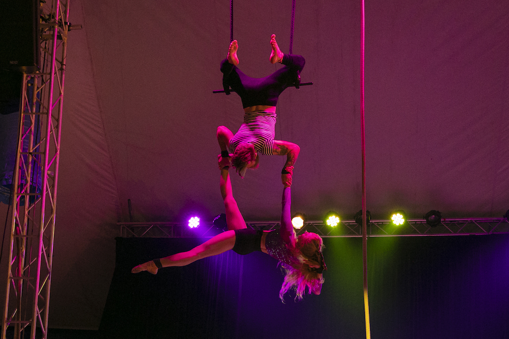 Two circus performers hang above a stage lit in pink and purple shades. One is hanging upside down, feet looped in the rings of a trapeze. This performer is holding the second horizontally by a leg and arm,
These are performers from the Kaleido Company, who are part of Fringe World.