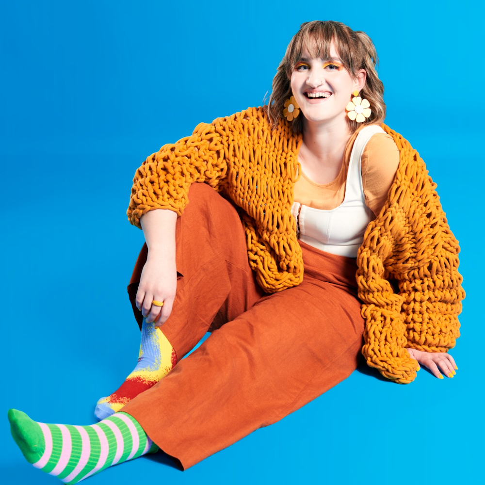 A woman wearing flower drop earrings and bright orange pants and knitted cardi over a white top sits on a blue background, her feet sticking out in odd socks. 
This is performer Gabbi Bolt, who brings her Odd Socks show to Fringe World.