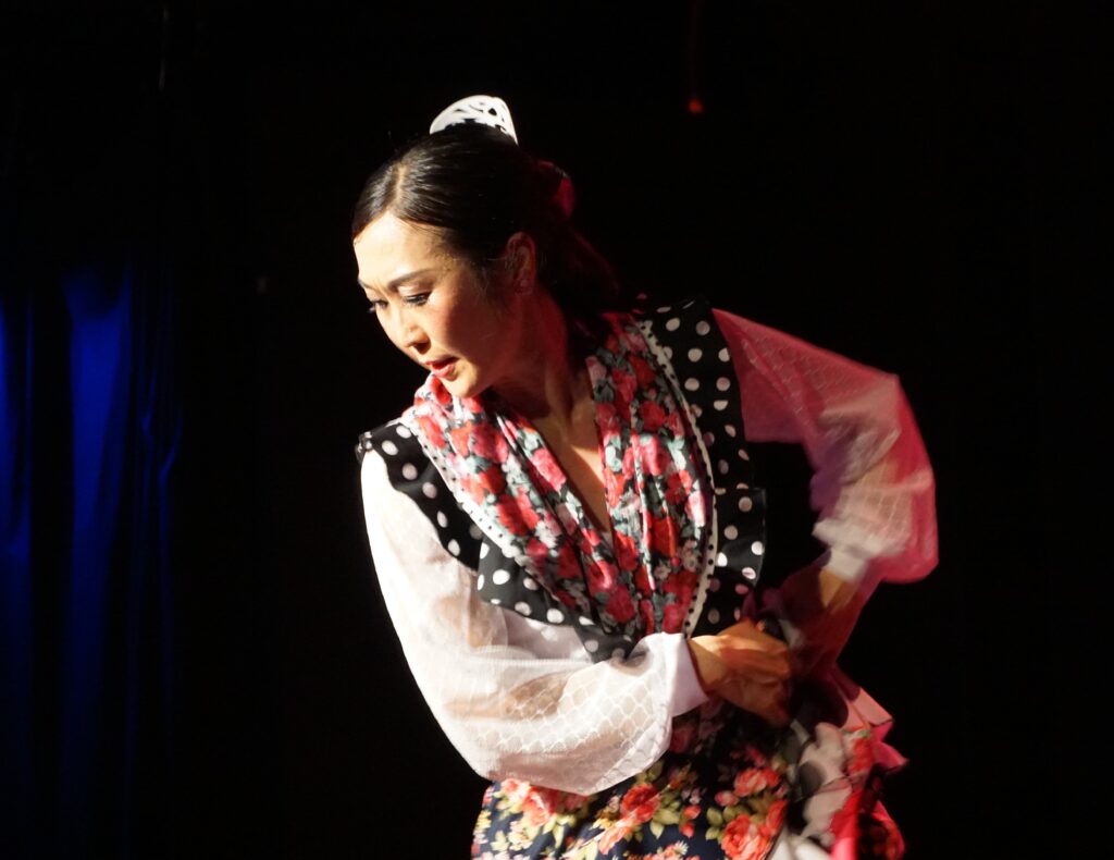 A dancer in a traditional costume strikes a pose. She her hair tied tightly back, shiny comb in her hair, flamenco style.
This is Naoko Christofis from La Pandilla Flamenca