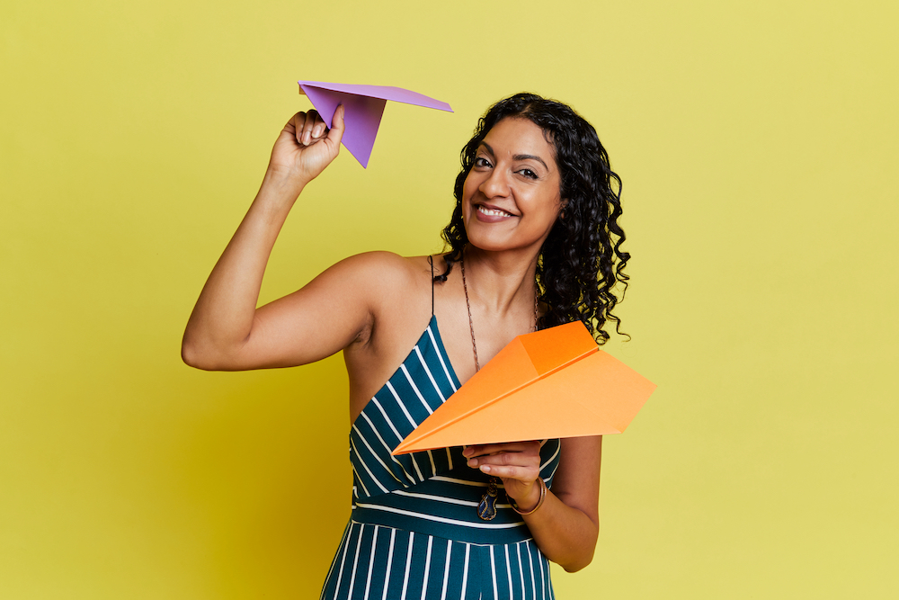 A smiling woman in a striped dress stands against a yellow backdrop. She is holding two paper planes, a purple one in one hand at head height, an orange one in front of her.
This is Canadian comedian Aliya Kanani, who is bringing her show to Fringe World.