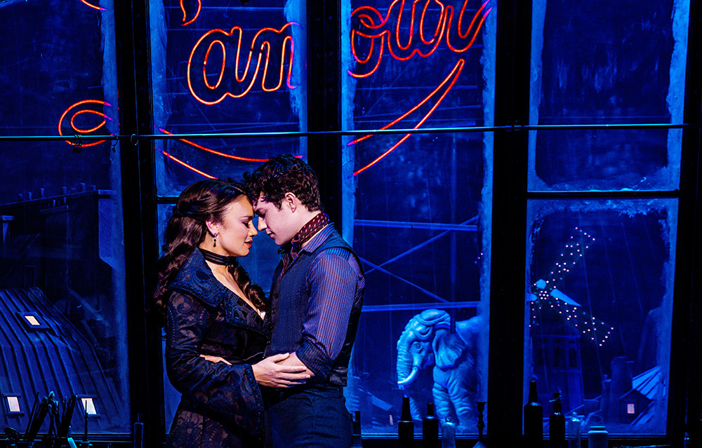 Alinta Chidzey as Satine and Des Flanagan as Christian holding each other on stage in 'Moulin Rouge! The Musical'. They are infront of windows and a sign which says Lamour. 
