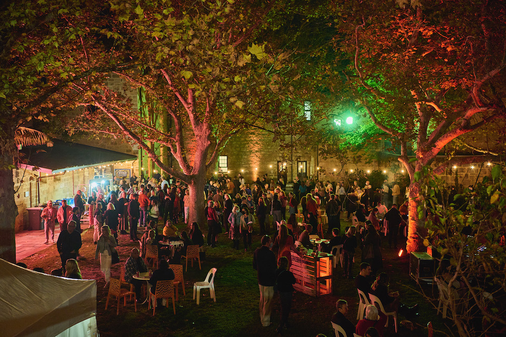 A large group of people are gathered under trees in a courtyard. The trees are bathed in green and red light, the building behind them cast in a golden glow. This is the Writers Weekend All the Stars Party.