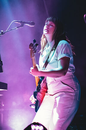 A woman wearing a blue top and pink skirt holds a guitar, singing plaintively into a fixed microphone. This is Wye Oak's Jenn Wasner at Bon Iver's concert.