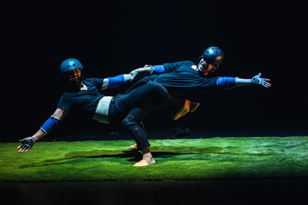 Two crash helmet-wearing men, one supporting the other around his waist, perform an acrobatic dance routine.